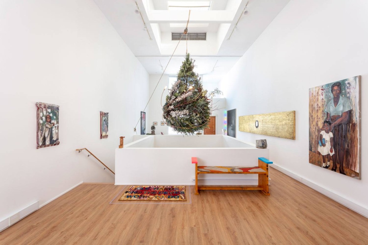 Art gallery interior with colorful paintings on white walls, wooden flooring, and a large hanging installation in the center.