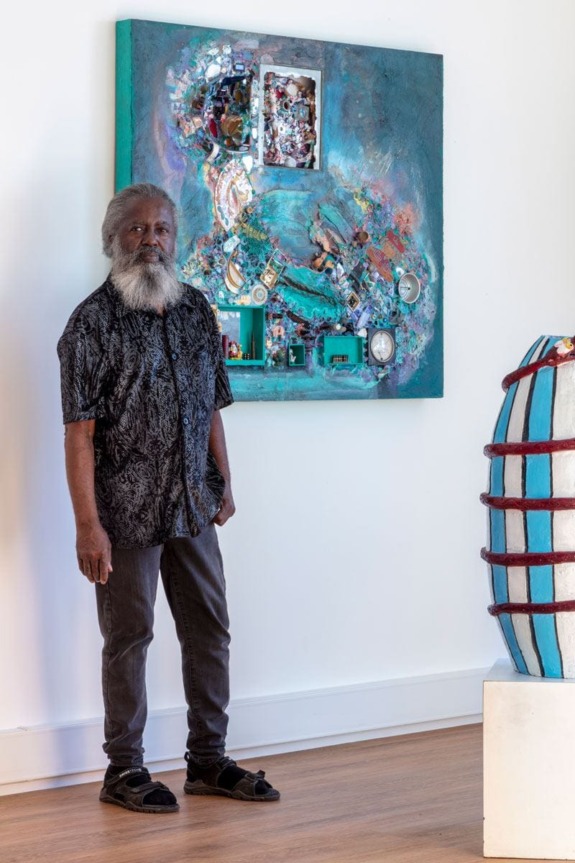 Man standing in art gallery next to a colorful abstract painting on the wall and a sculpture on the floor.