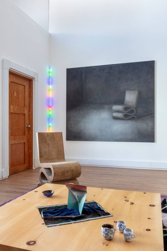 Modern interior with a large painting of a chair, a wavy chair, colorful lights, a wooden door, and a wooden table with decor.