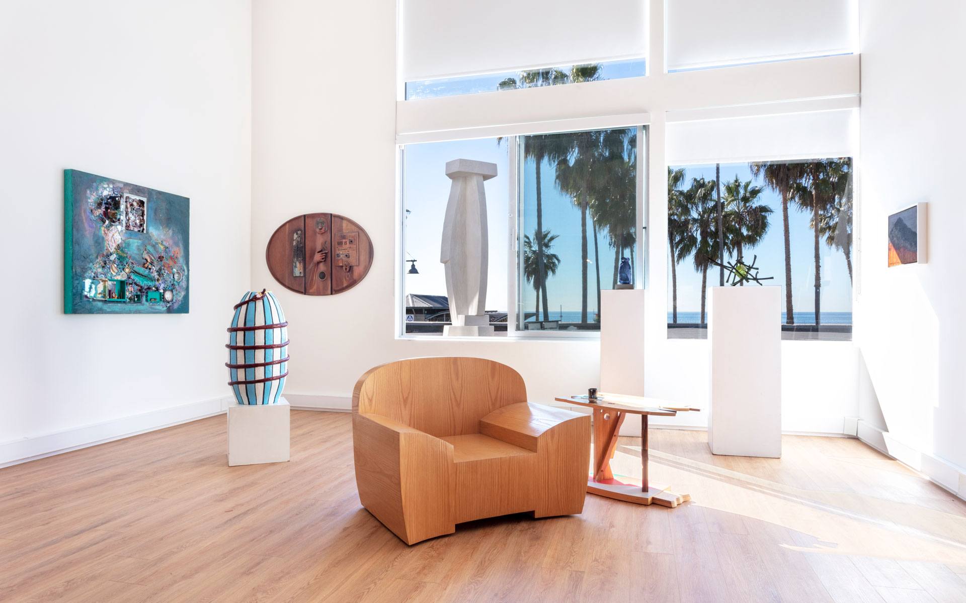 Modern art gallery with wooden chair, vase, paintings, and large windows showing palm trees outside.