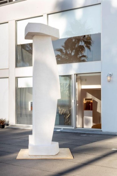 White abstract sculpture displayed outdoors in front of a modern building with large windows and palm tree reflections.