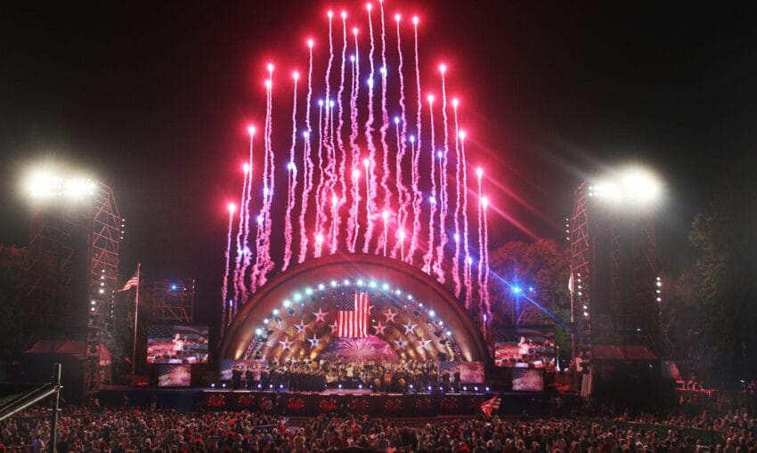 Fireworks display over a concert stage with large crowd at night, featuring an American flag backdrop.