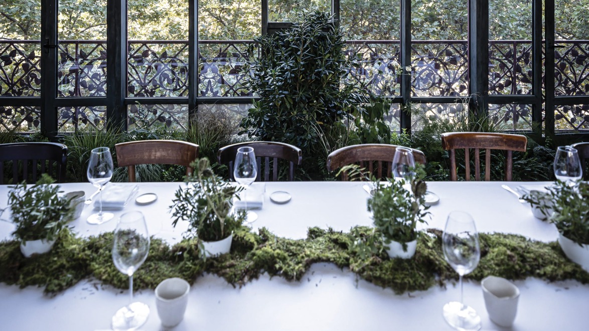 Elegant dining table set with greenery, glassware, and cutlery in a sunlit room with large windows and garden view.