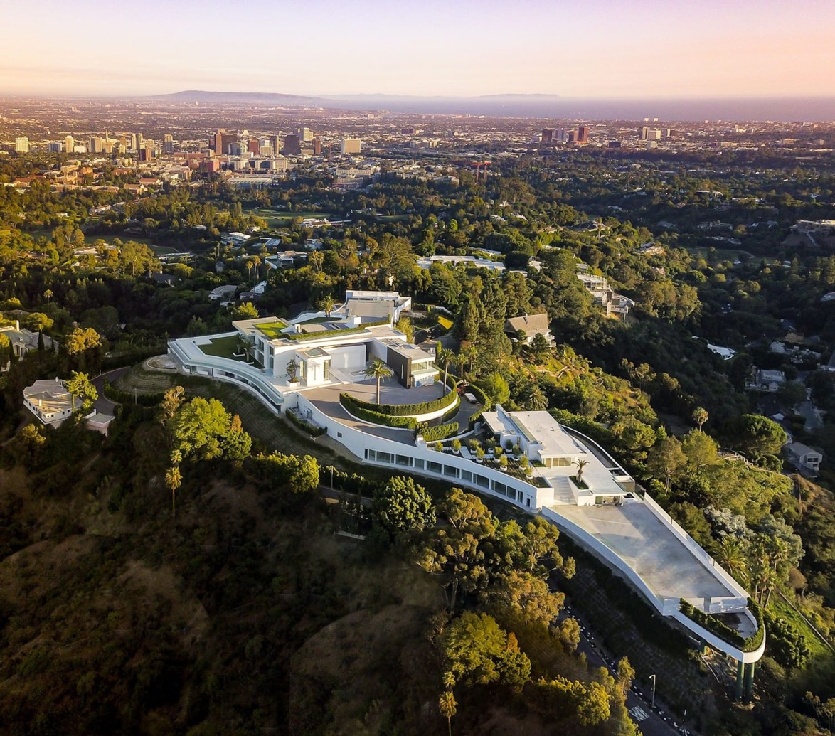 Aerial view of a large, luxurious mansion nestled in greenery on a hill with a cityscape in the background under a clear sky.