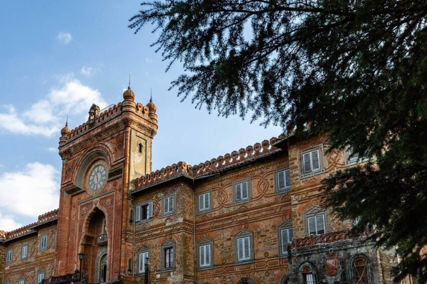 Historic brick building with ornate details and a large clock tower, framed by tree branches under a blue sky.