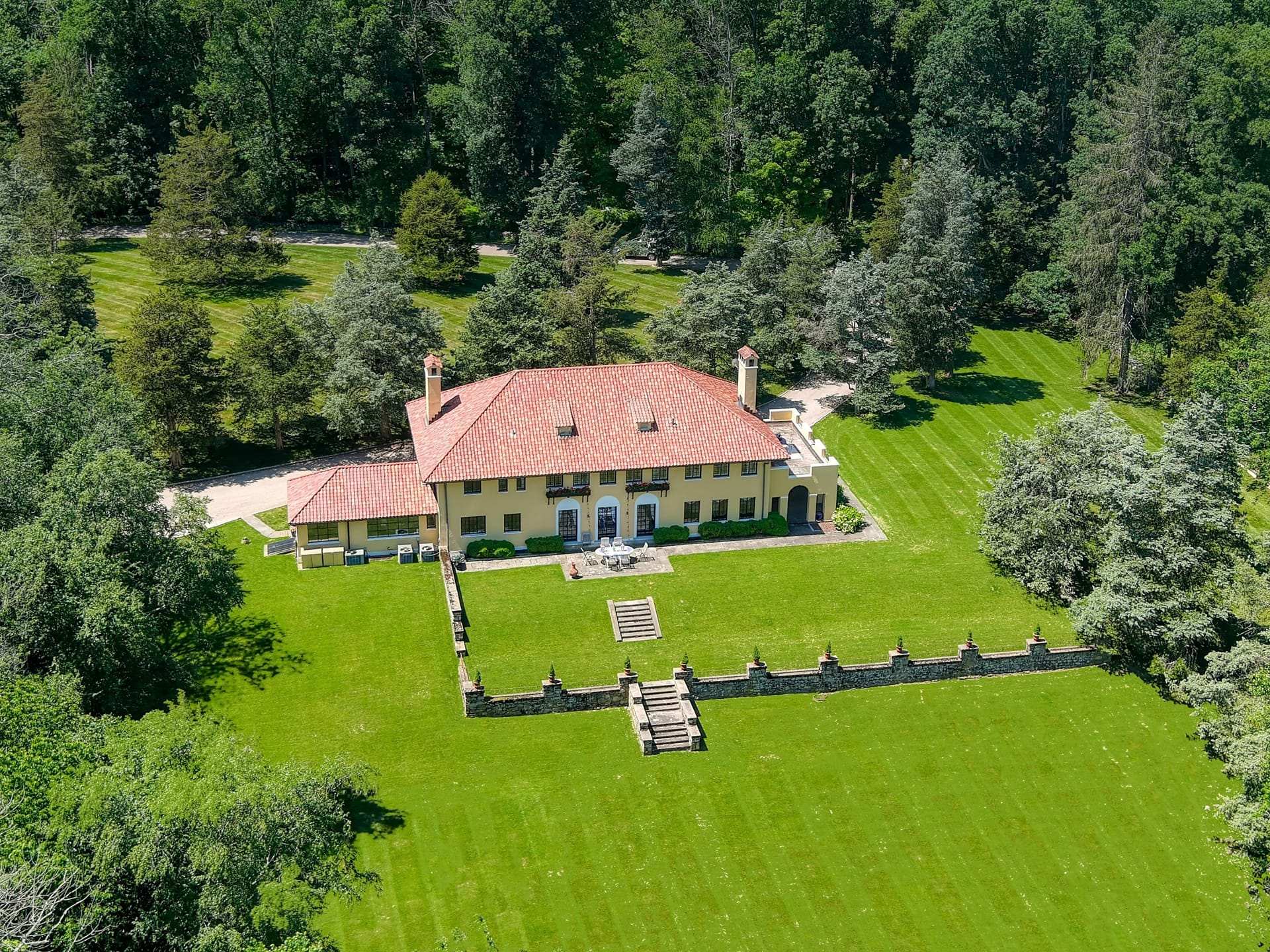Aerial view of large mansion with red roof and spacious green lawn surrounded by trees.
