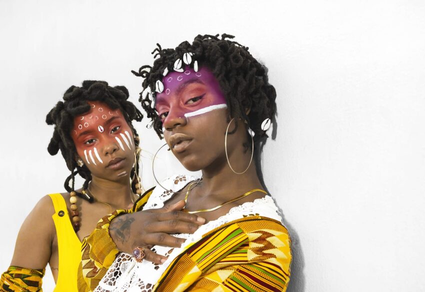 Two individuals with intricate face paint and traditional attire pose confidently against a plain background.