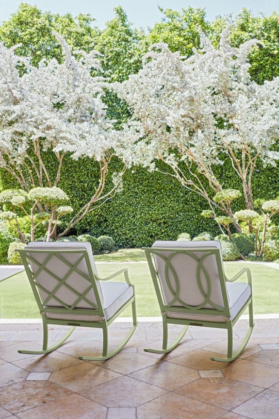 Two white patio chairs facing a lush garden with neatly trimmed hedges and blooming trees on a sunny day.