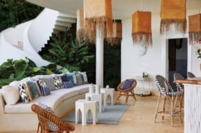 Outdoor patio with woven chairs, tables, and decorative pillows, surrounded by lush greenery and spiral staircase in background.