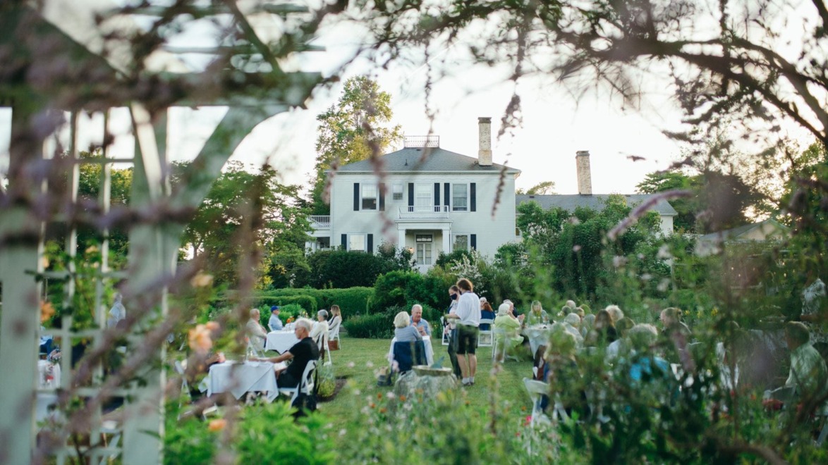 Outdoor wedding reception with guests seated at tables in a garden setting with a large house in the background.