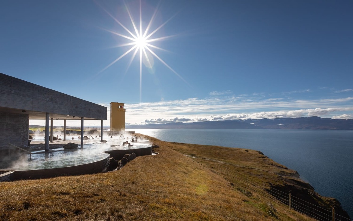 Sunlit geothermal spa by a scenic cliffside overlooking the ocean with clear blue skies and mountains in the background