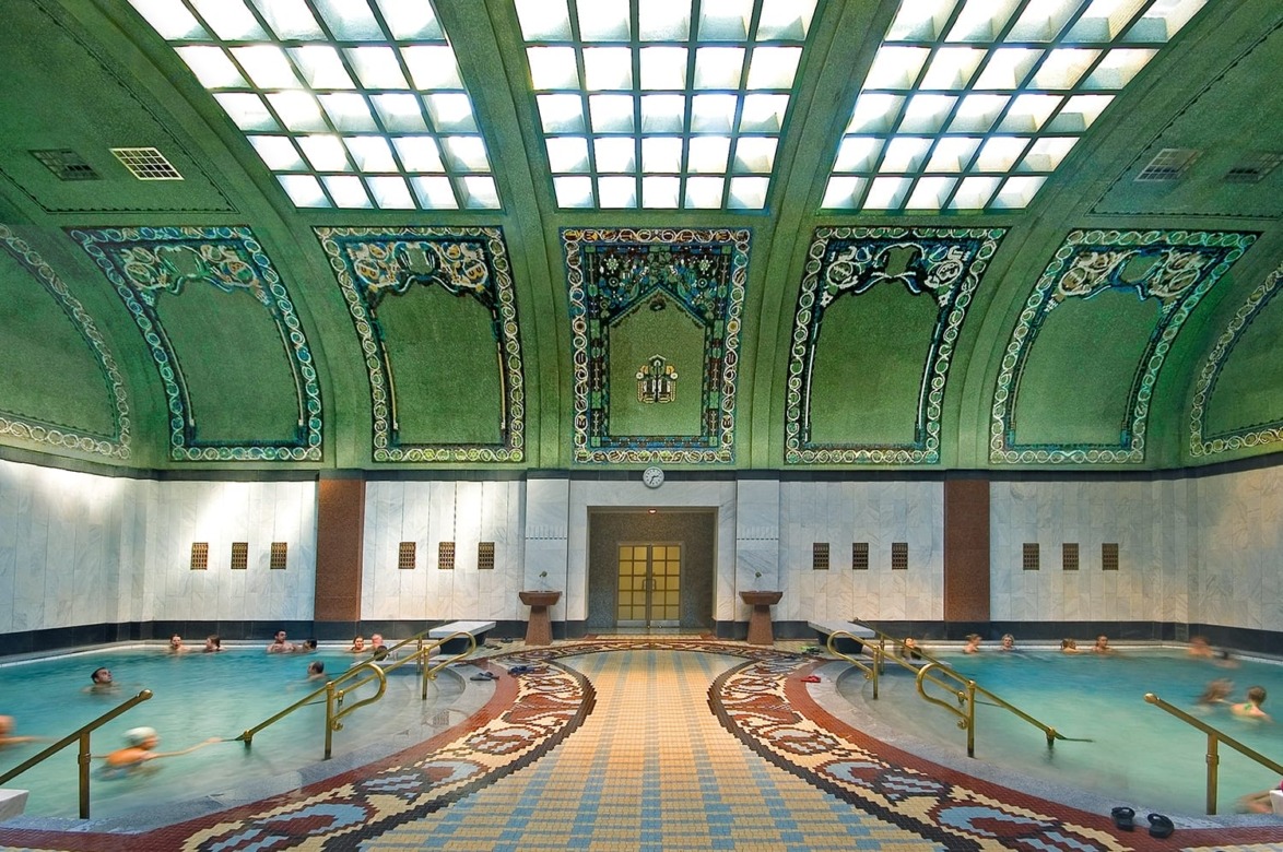 People relaxing in an ornate indoor pool with a decorative tiled floor and stained glass ceiling.