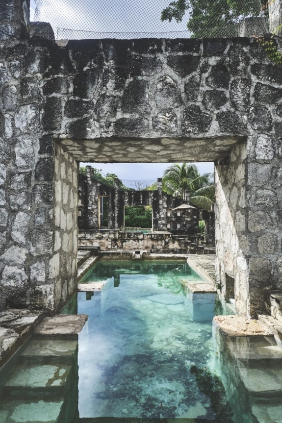 Ancient stone ruins with clear blue water reflecting the sky, surrounded by lush greenery and palm trees.