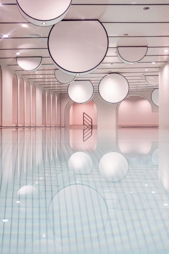Spacious modern indoor swimming pool with mirrored ceiling, circular lights, and elegant archways reflecting in the water.
