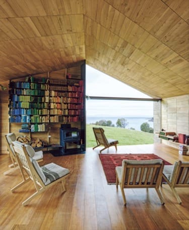 Modern wooden interior with colorful bookshelves, chairs, and a window view of a green landscape and water.