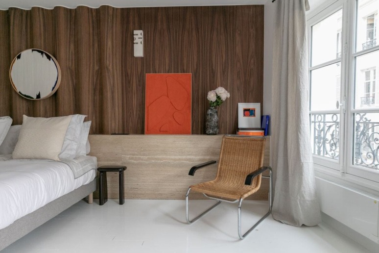 Modern bedroom with wood-paneled wall, abstract art, wicker chair, and a large window with gray curtains.