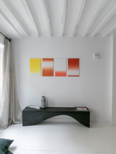 Minimalist room with white walls, abstract art, dark wooden bench, a vase, and rolled-up magazine, natural light through window.