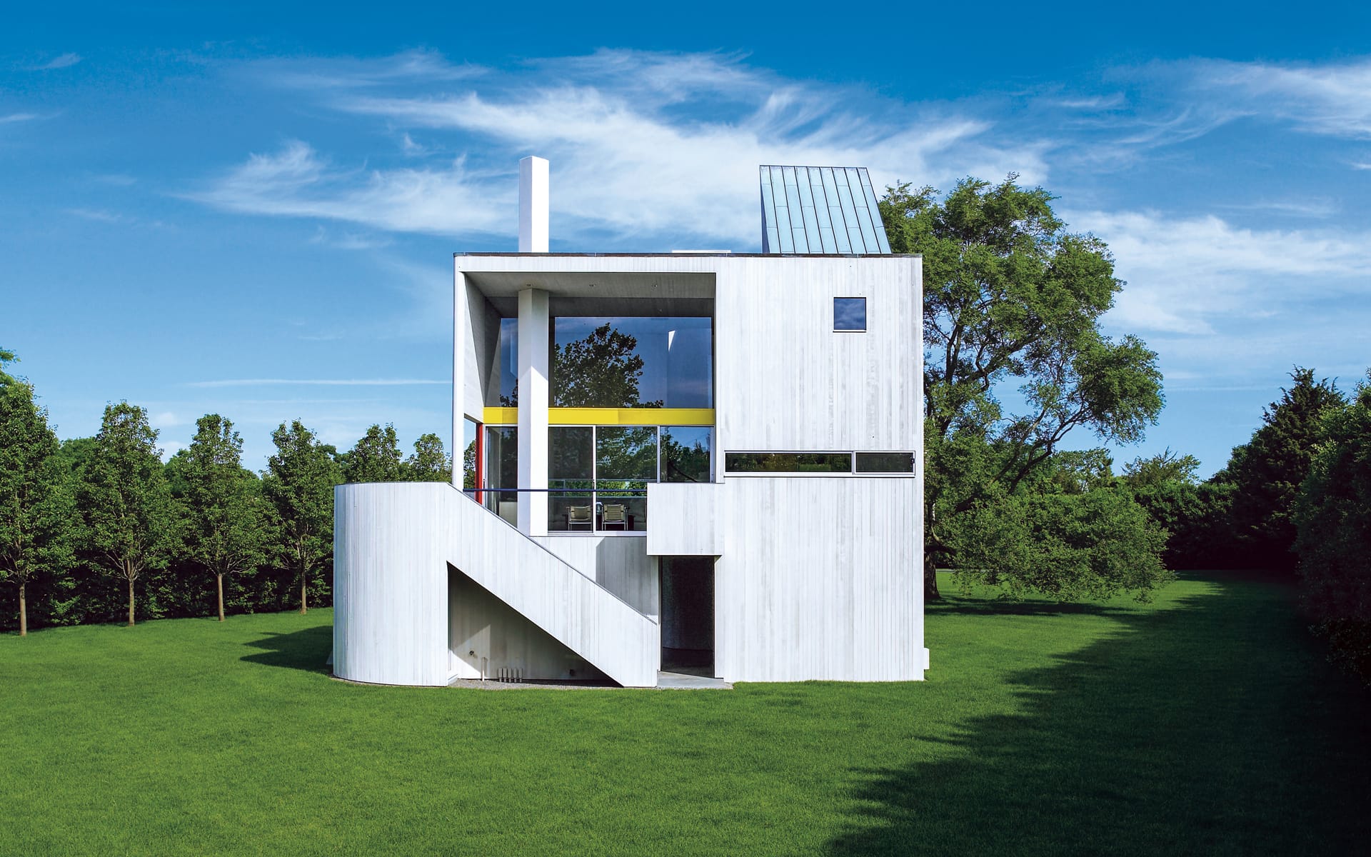 Modern white house with large windows, surrounded by green grass and trees under a clear blue sky.