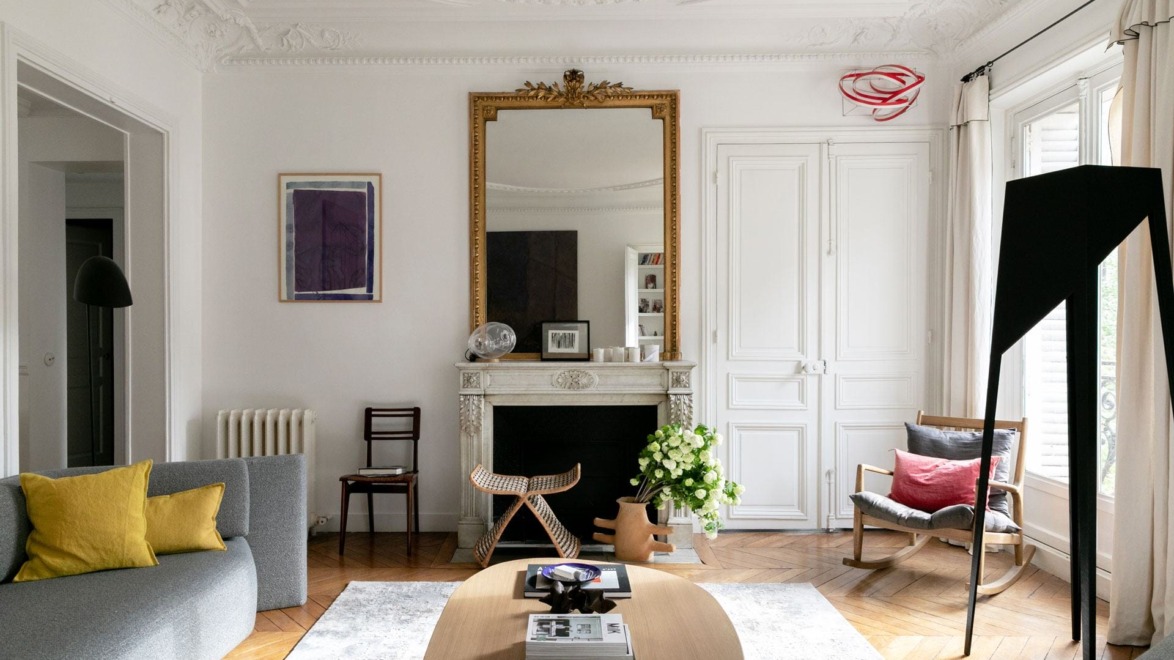 Elegant living room with ornate ceiling, large mirror above a fireplace, modern furnishings, and natural light from tall windows.