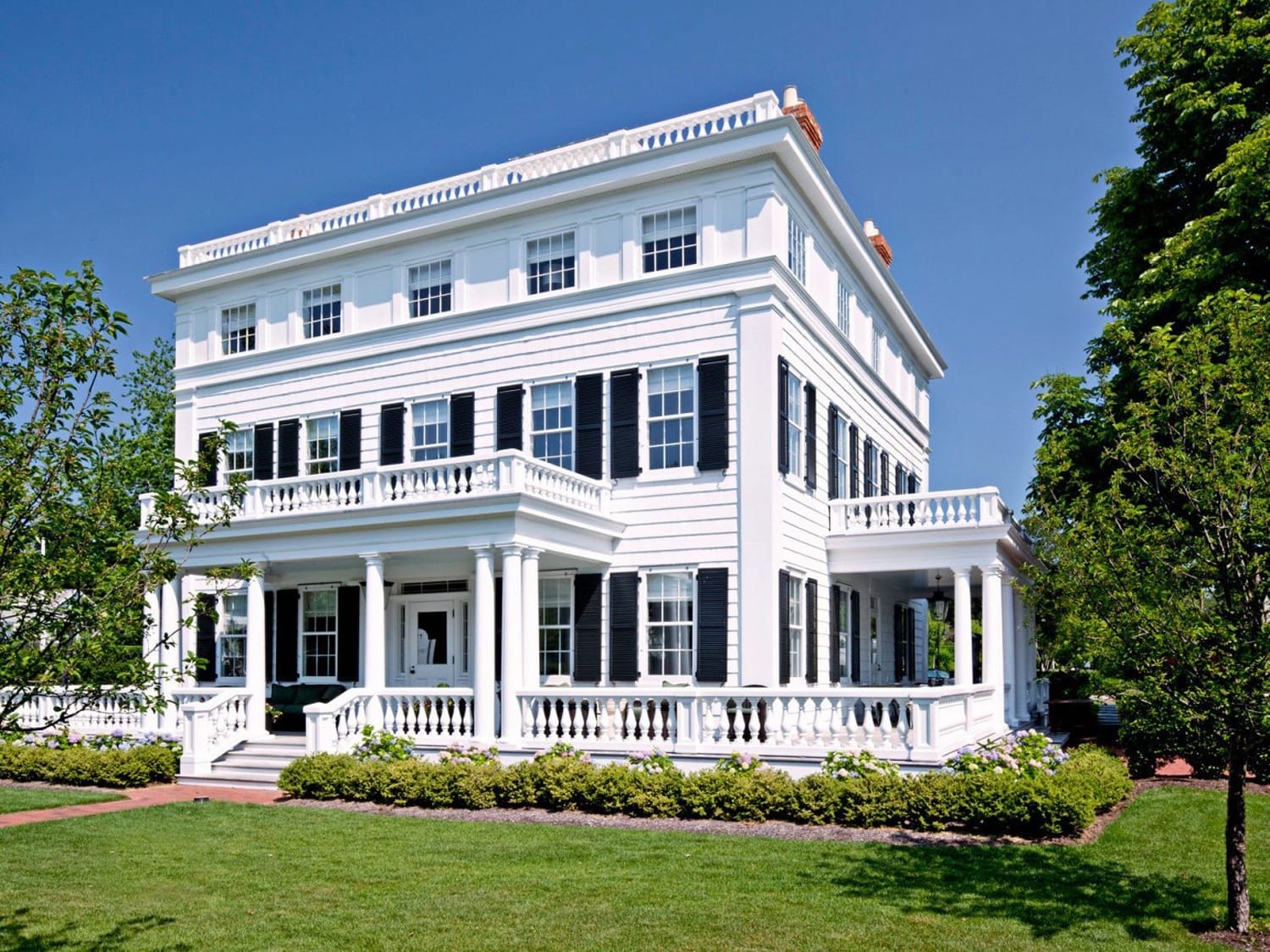 Elegant white colonial-style house with black shutters, surrounded by green lawn and trees under a clear blue sky.