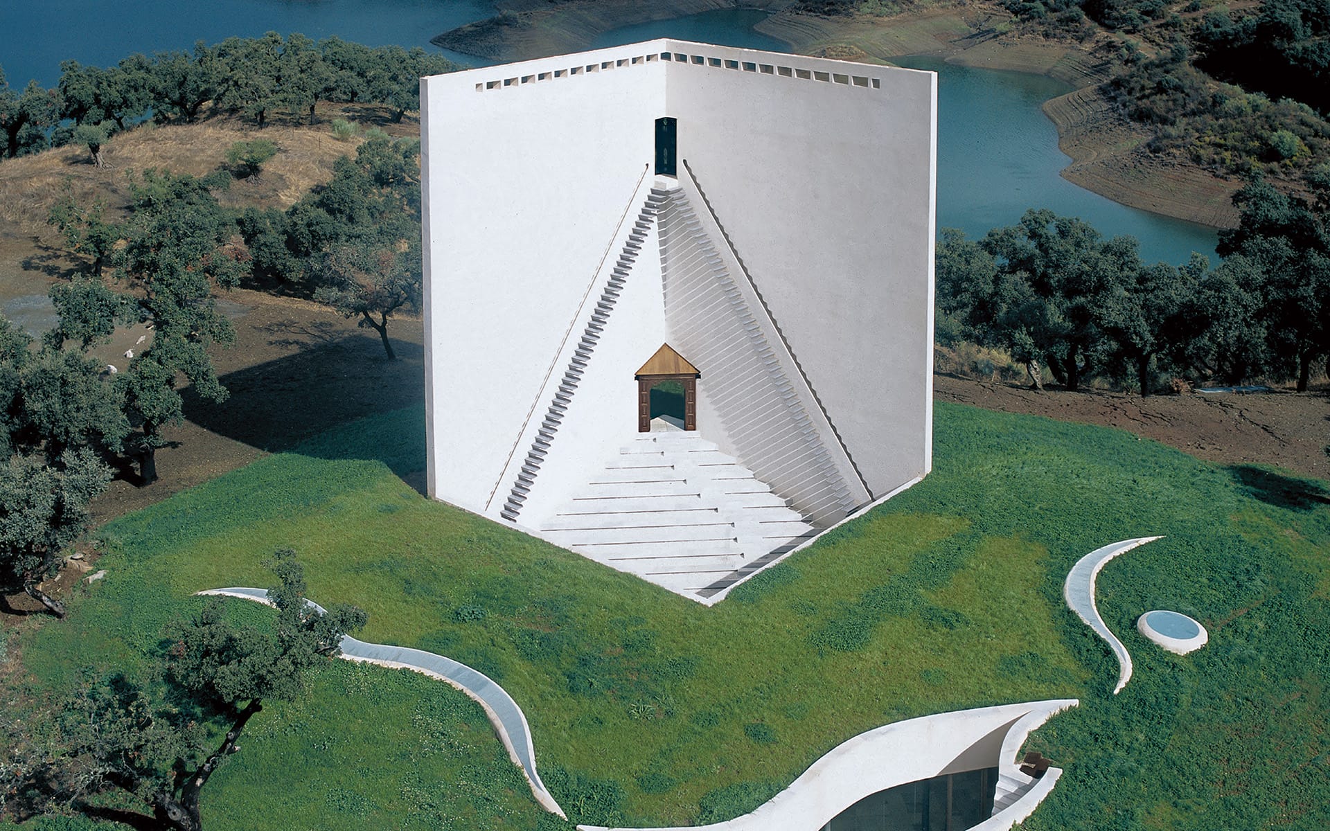 White geometric building with triangular steps on a grassy landscape, surrounded by trees and water in the background.