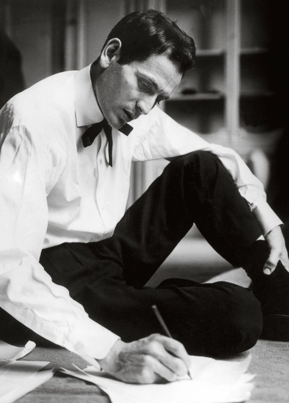 Man sitting on floor, wearing a bow tie and writing with a pen on papers spread out in front of him.