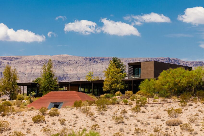 Modern desert house with trees and mountains in the background under a bright blue sky with scattered clouds.