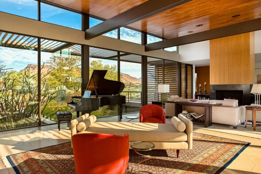 Modern living room with large windows, grand piano, beige sofa, red chairs, and scenic mountain views in the background.