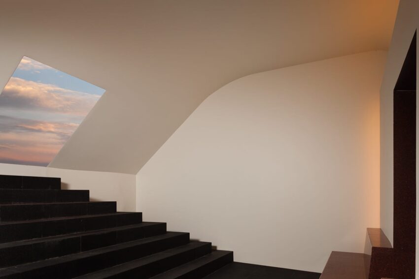 Modern interior with black staircase, slanted white ceiling, rectangular window showing a cloudy sky with a hint of sunset colors.