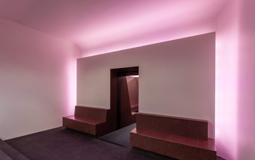 A modern interior with pink lighting, featuring minimalist benches and an angular dark entrance in the center.