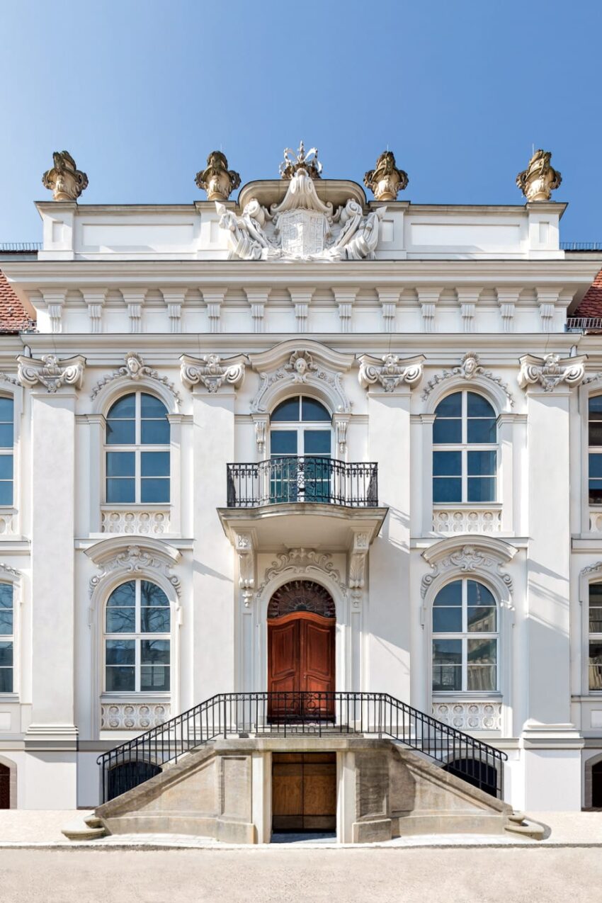 Ornate white baroque building facade with decorative sculptures, arched windows, and a grand entrance with staircase.