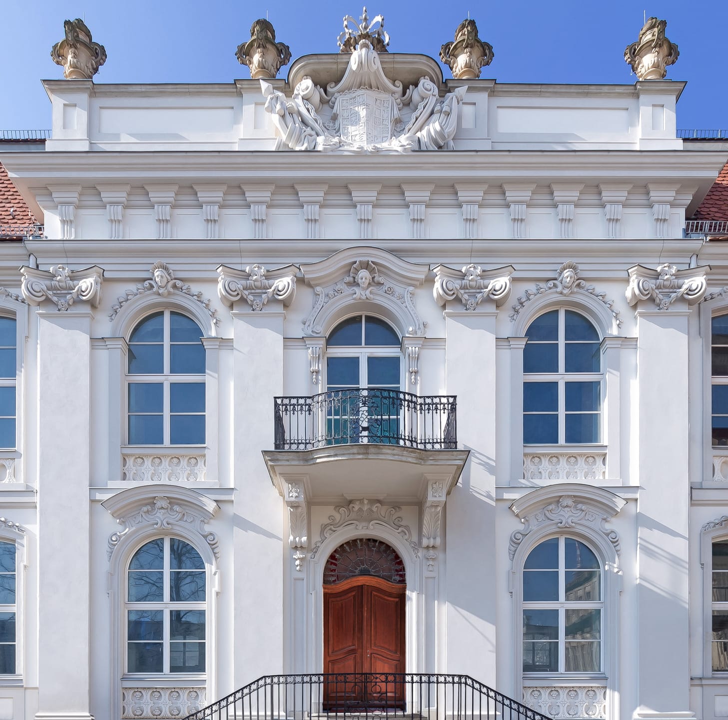 Ornate facade of a historic white building with arched windows, a central balcony, and intricate sculptures under a clear blue sky.