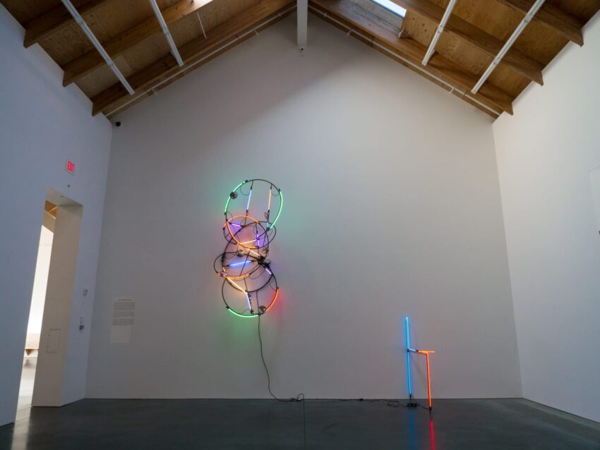 Art installation in a gallery with colorful neon lights on the wall and a neon light chair on the floor.
