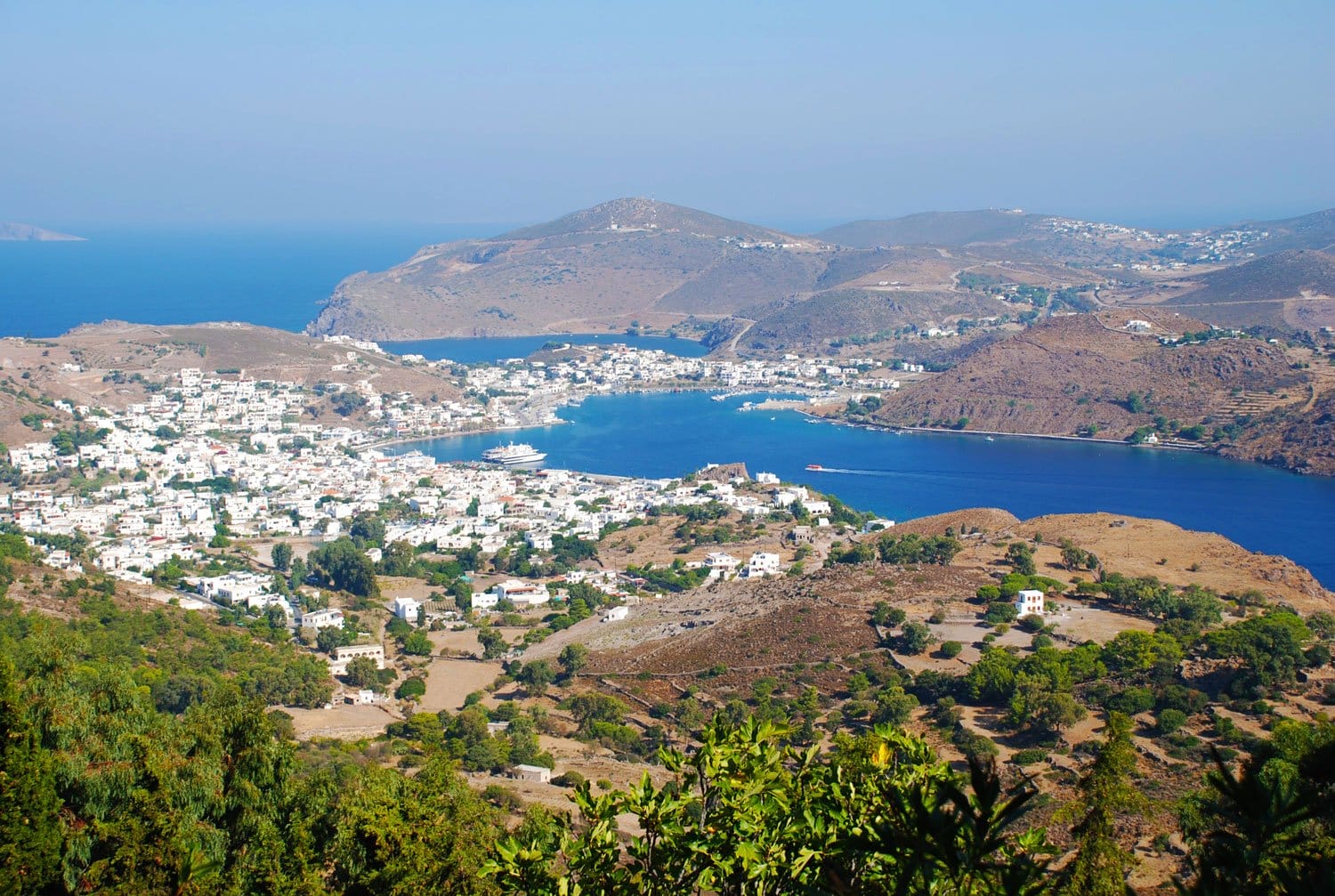 Aerial view of a coastal town with white buildings, blue sea, and mountainous landscape in the background.