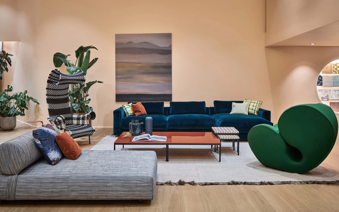 Modern living room with blue velvet sofa, abstract painting, and various stylish chairs on a light wood floor.