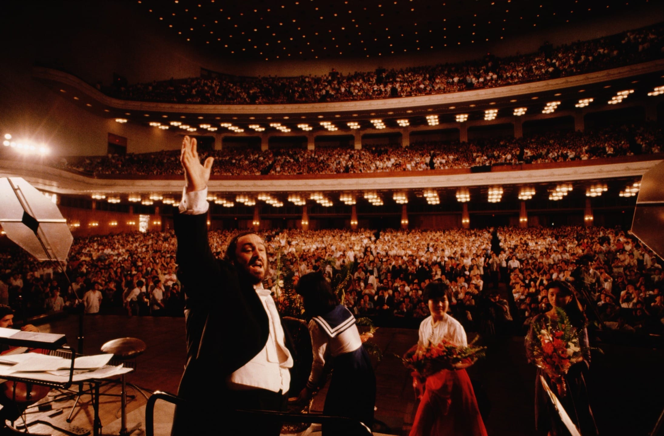 Opera singer on stage waving to a large audience in a grand theater, with people holding flowers in the foreground.