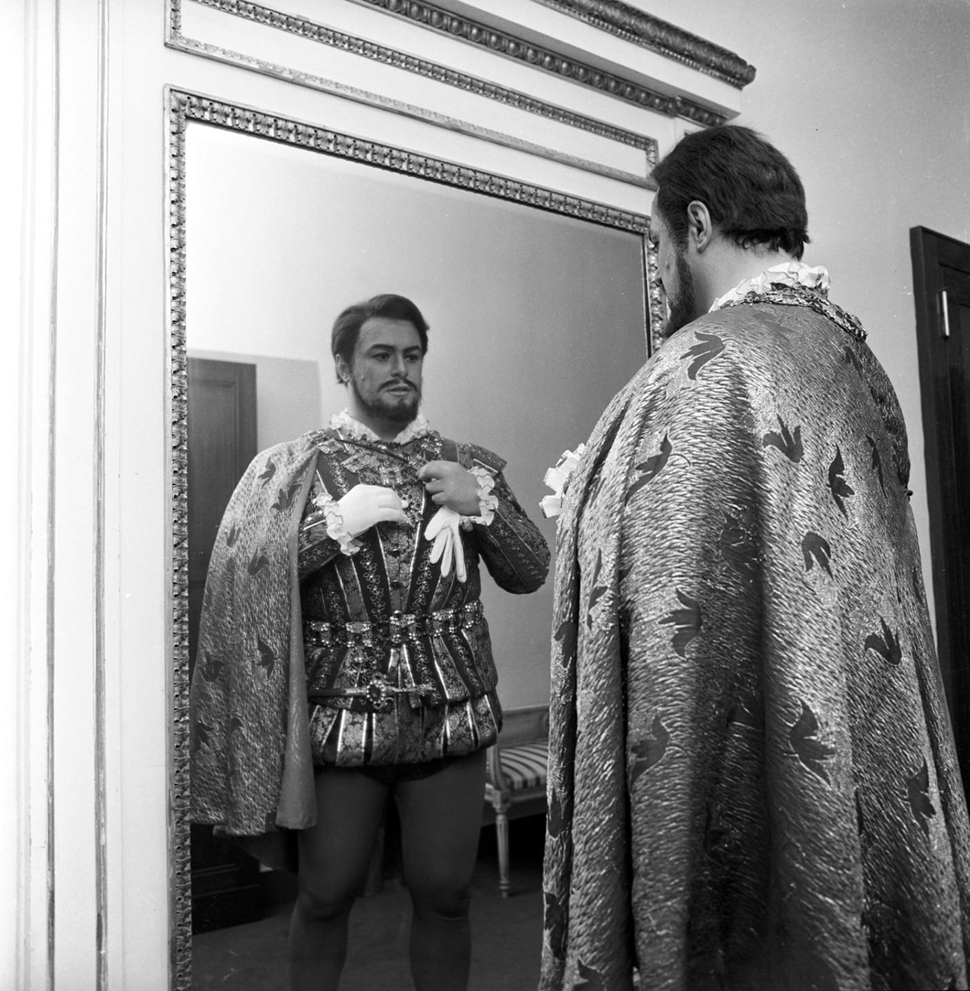 Man in an elaborate costume looking at himself in a large ornate mirror, adjusting gloves in a dressing room setting.