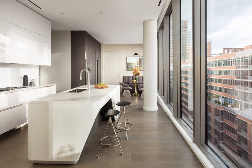 Modern apartment kitchen with sleek white island, bar stools, and large windows offering a cityscape view.