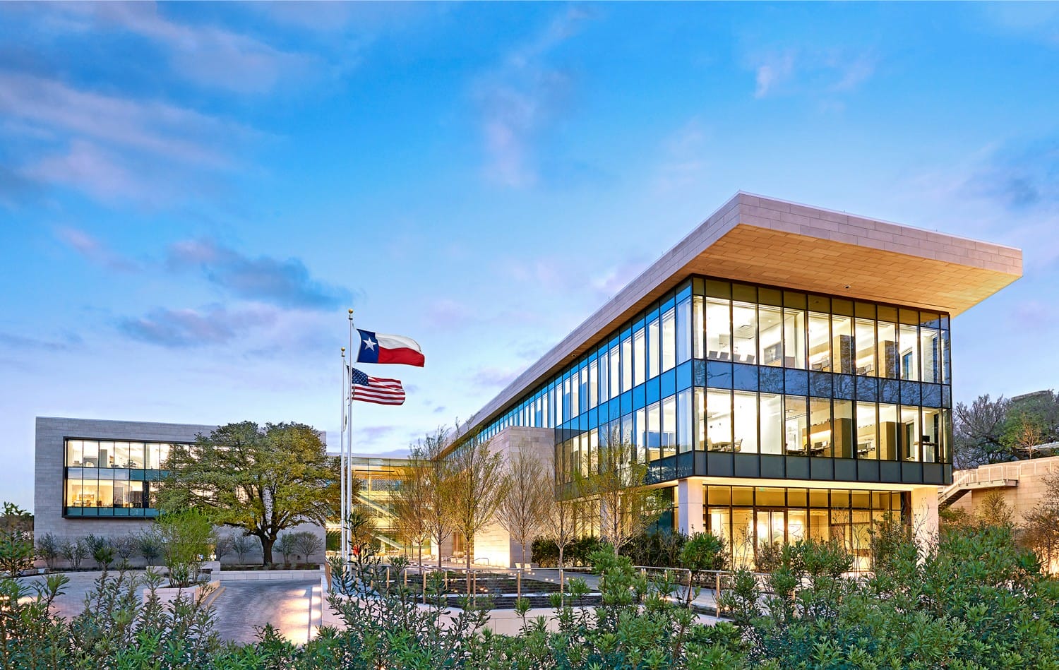 Modern building with glass facade at dusk, featuring a Texas flag in the foreground and surrounded by trees.