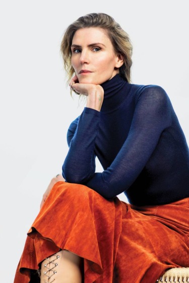 Person sitting in thoughtful pose, wearing a blue turtleneck and orange skirt against a light background.