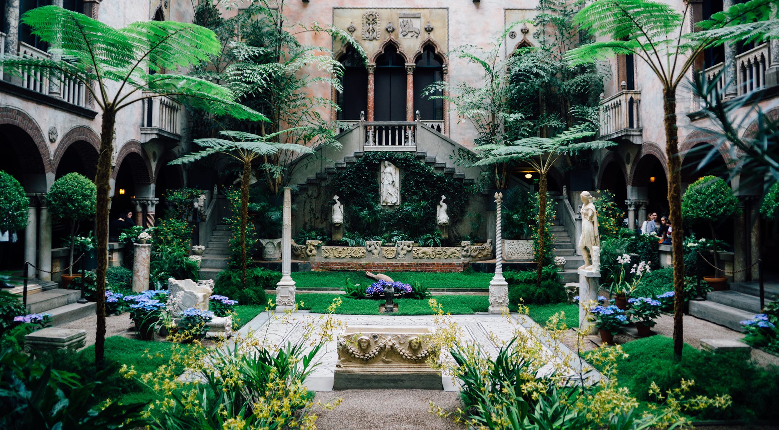 Lush courtyard with tall ferns, ornate stone staircase, sculptures, and vibrant greenery in a historic setting.