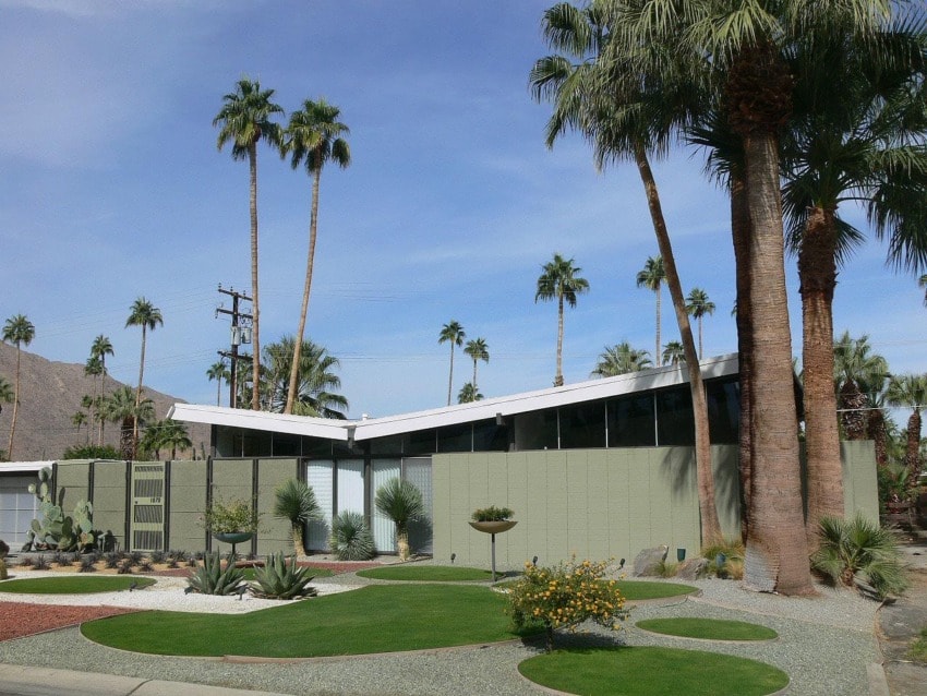 Mid-century modern house with angular roof, surrounded by palm trees and landscaped yard under a clear blue sky.
