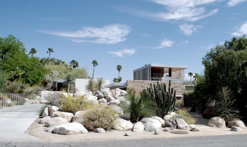 Modern desert home surrounded by rocks, cacti, and palm trees under a blue sky with wispy clouds.
