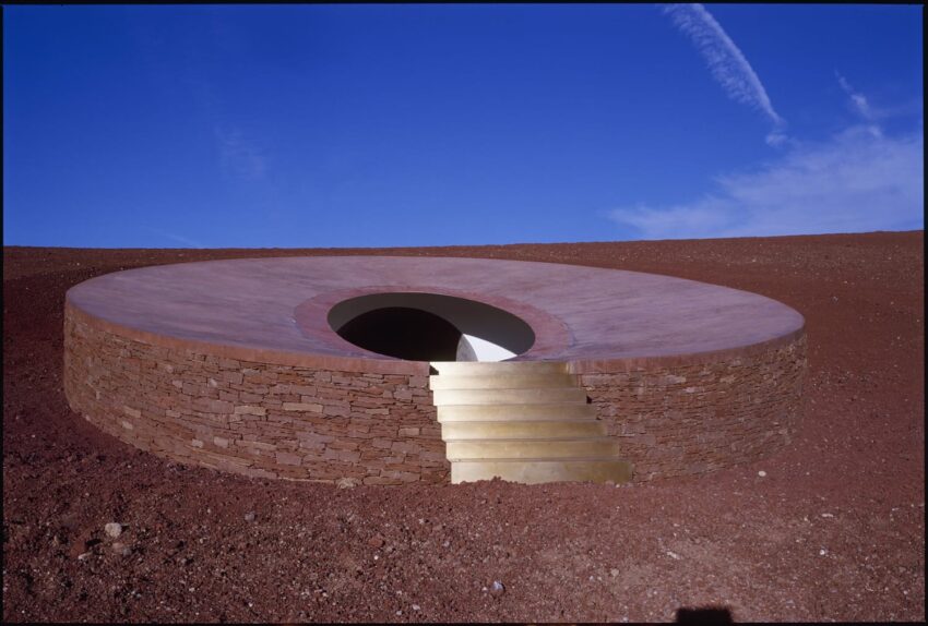 Circular stone structure with central opening and stairs, set in a red earth landscape under a clear blue sky.