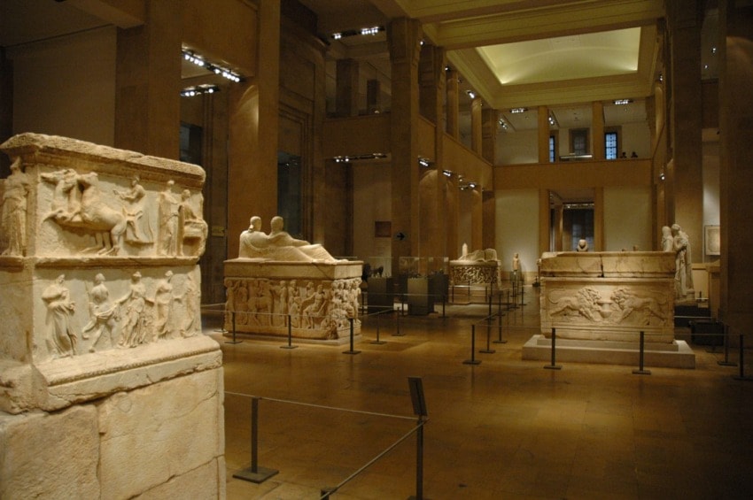 Museum gallery showcasing ancient stone sarcophagi and sculptures under soft lighting and high ceilings.