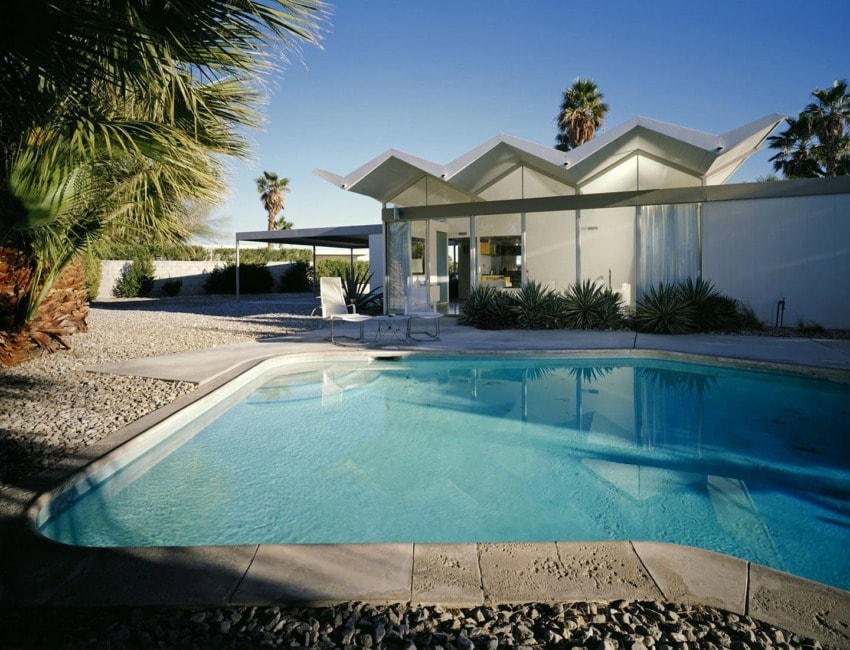 Modern house with geometric roof design, clear blue swimming pool, surrounded by palm trees and stone landscaping.