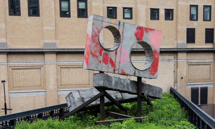 Modern outdoor sculpture with circular cutouts, featuring red and gray abstract patterns, surrounded by greenery.
