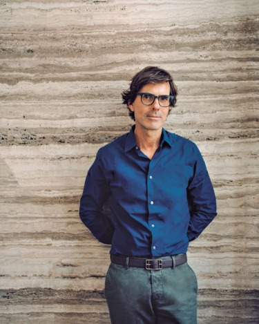 Man in blue shirt standing against a stone-textured wall with arms relaxed behind his back.
