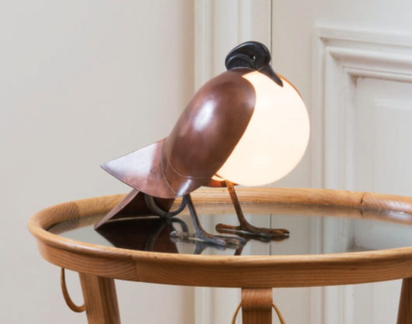 Bird-shaped lamp on a round wooden table, designed with metal and glass, illuminating a cozy room corner.