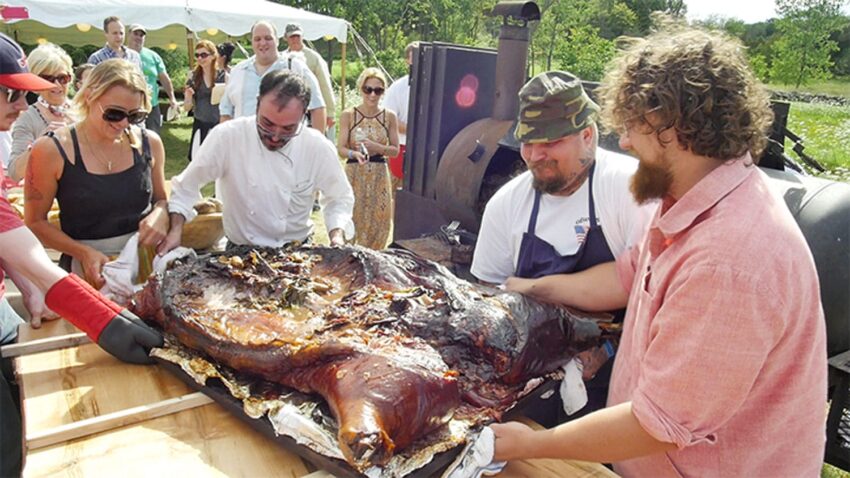 Group of people preparing a roasted whole pig outdoors near a barbecue smoker.